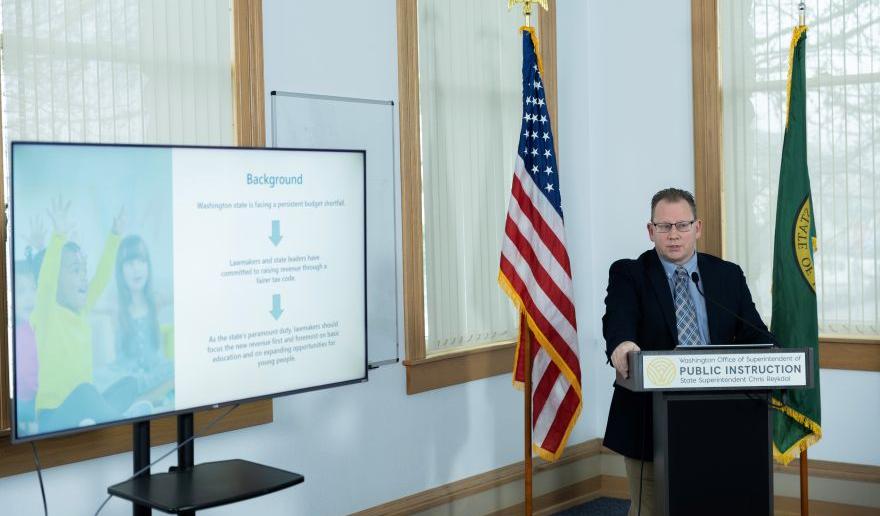Superintendent Reykdal behind a podium next to a T.V. screen 