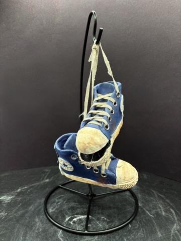 A pair of small, worn blue sneakers with white laces and soles are hanging by their laces from a black metal stand. The sneakers appear to be old and used, with visible dirt and scuff marks. The background is dark, highlighting the shoes as the main focus of the image.