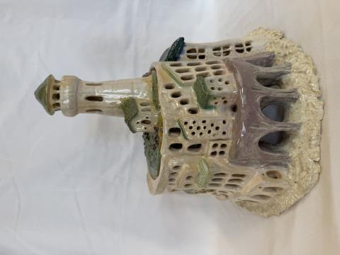 A ceramic model of a castle with intricate details, including multiple windows, arches, and a tall tower. The castle is painted in shades of beige, green, and brown and sits on a textured base that resembles rocky terrain.