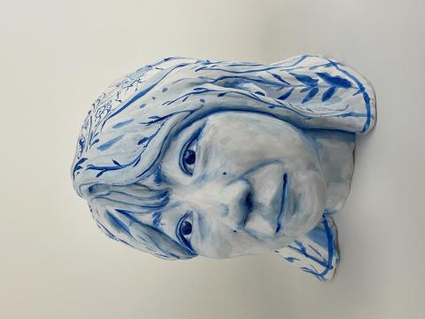 A ceramic bust of a person's head and shoulders, painted in shades of blue and white. The face has a calm expression with detailed features, and the hair is adorned with intricate blue patterns resembling leaves and branches.