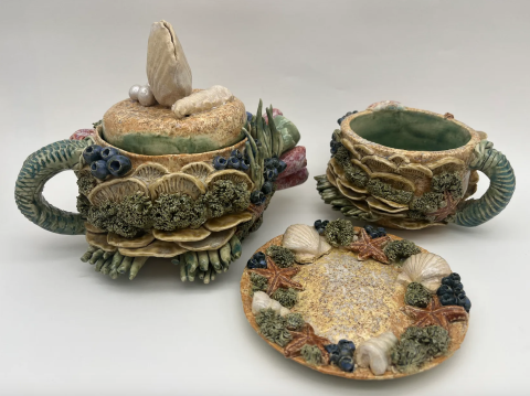 A set of intricately designed ceramic tableware, including a teapot with a lid, a cup, and a saucer. The teapot features detailed sea-themed decorations such as seashells, starfish, and coral-like textures.