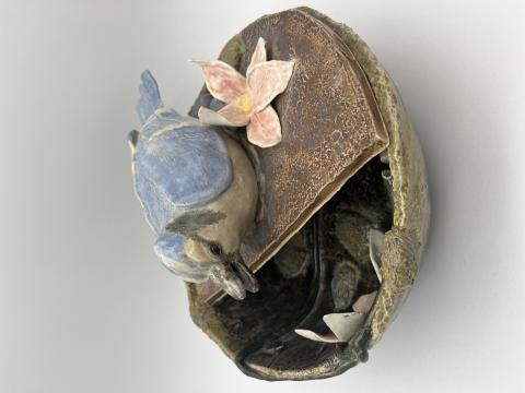 A ceramic sculpture of a bluebird perched on the edge of a rustic, round container. The bird is detailed with blue and beige feathers and has its beak slightly open.