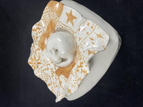 A ceramic sculpture featuring a central, smooth, white animal surrounded by an intricately patterned, orange and white square base with wavy edges. The base has various geometric and floral designs