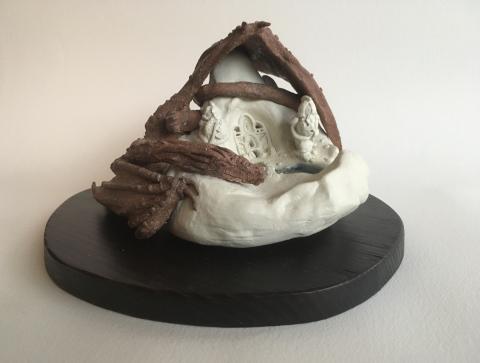 A small, intricate sculpture placed on a dark oval base. The sculpture appears to be made of clay and depicts a scene with detailed figures and structures inside what looks like a cave or grotto.