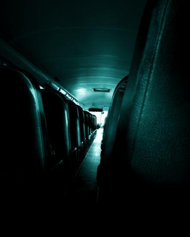 The interior of an empty bus, viewed from the back towards the front. The seats are arranged in rows on either side of a central aisle. The lighting is dim, casting a blue-green hue over the scene.