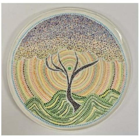 A circular artwork featuring a tree at the center, created using a pointillism technique. The tree has a black trunk and branches, with foliage depicted in various shades of blue, purple, and white dots. 