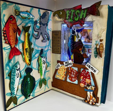 An open book creatively transformed into a 3D art piece. The left page features colorful fish illustrations, each uniquely designed with different patterns and colors. The right page depicts a small fish market scene with a wooden stall displaying labeled sections for "Cod," "Red Snapper," and "Salmon." Above the stall, there is a sign shaped like a fish with the word "FISH" on it. 