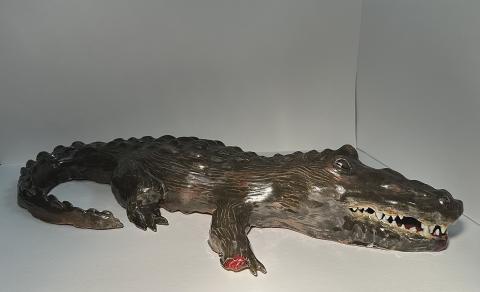The artwork is a realistic sculpture of a crocodile displayed against a plain background. The sculpture is detailed, showing the textured skin, sharp teeth, and claws of the crocodile. 
