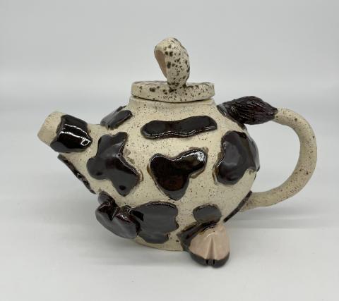 The artwork is a ceramic teapot designed to resemble a cow. The teapot is primarily white with black spots, mimicking the pattern of a cow's hide. The spout and handle are shaped like a cow's head and tail, respectively. 