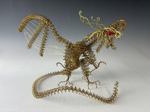 The image shows a detailed sculpture of a dragon made from intricately twisted and coiled wire. The dragon has large wings, a long tail, and an expressive face with what appears to be a red bead for an eye. 