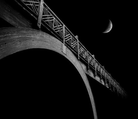 A digital photograph of a bridge against a black night sky with a crescent moon above