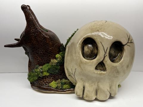 This photo displays a large brown speckled ceramic snail, with a faded skull chapped shell. The whole piece is lined with different shades of green moss.