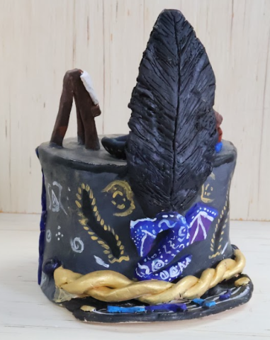 A marching band hat with a grey base, geometric designs in white, gold, and black across it. The brim has music notes and a golden braided band. The back of the hat has blue waves with cracks painted gold and the front of the hat has a dragon and black plume. The top has multiple small items, books, a bowl and a lotus flower next to an easel.