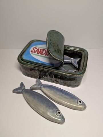 A life-size ceramic version of a sardines can with its lid being pulled back to find four individual clay sardines inside. The can has a dark gray and greenish color to it, while the lid of the can has a colorful label with red lettering. All four sardines have this blue and gray ombre glaze effect and have big, animated eyes.