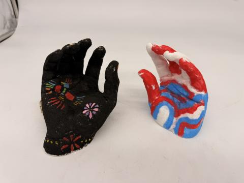 two hands, one red, white, and blue to represent USA which is posed as if it is sinking. Other hand is Black with Hispanic heritage designs on it reaching out as if to grab USA hand.