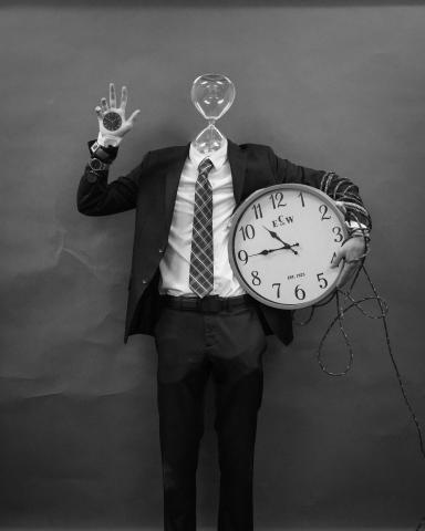 A black-and-white photograph of a suited man with his head replaced by an hourglass. Wristwatches cover one arm and a clock face is embedded in his raised palm. His other arm holds a large wall clock and is wrapped in ropes that trail out of the frame.
