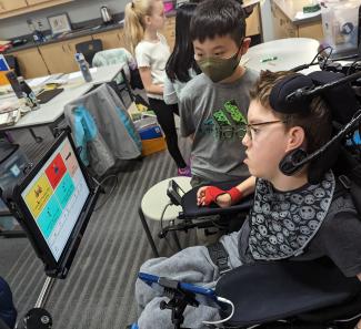 Jacob, sitting in his wheelchair and looking towards his AAC device, telling his classmate, “I would choose the same as you” from a choice of four options. Jacob is wearing gray sweatpants and tee shirt, and a gray neck scarf with animations on it. He has short brown hair and glasses. His peer, standing next to him and looking at Jacob’s AAC screen, is wearing a gray tee shirt and an olive-colored mask, and has short black hair. There are other students in the background, also standing together talking.