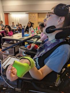 Jacob, a 5th grader who is using an AAC device attached to his electric wheelchair, is participating in his general education classroom large group discussion. He has short brown hair, and is wearing glasses. He has on a blue tee-shirt, a neck scarf with pineapples on it, has a green splint on his left hand, and a red hand splint on his right hand. In the background are Jacob’s peers waiting for Jacob’s response to the topic.