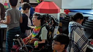 Jacob, dressed in his pajamas, is looking towards the front of the room alongside a peer.It is a little difficult to see Jacob as he is surrounded, in the best of ways, by his peers celebrating a pajama party together. He is wearing a rainbow neck scarf, has short brown hair, and glasses.