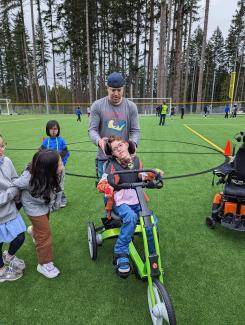 Jacob is using a three-wheeled green bike out in the school soccer field. He is smiling and looking to the side at his peers who are running along by his side. A teacher is standing behind Jacob’s bike, adjusting his head rest. Jacob’s electric wheelchair is off to the side while he is biking. 