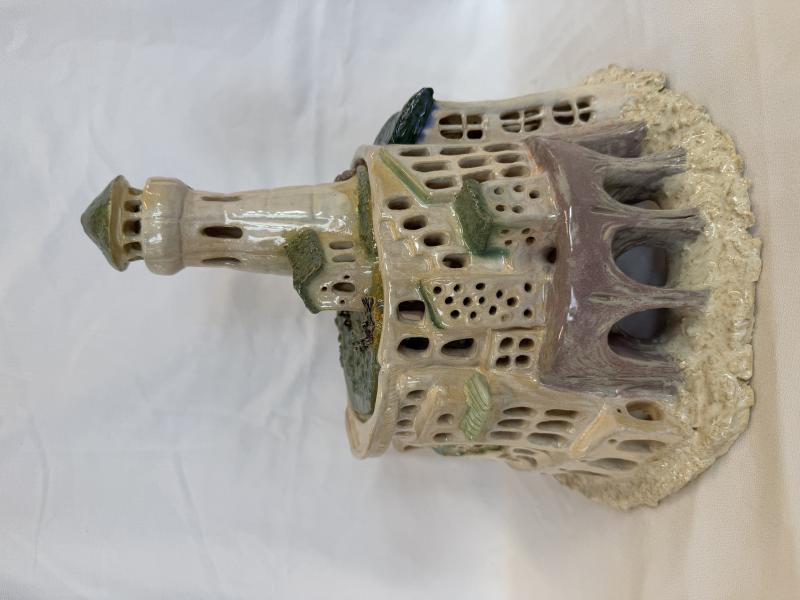 A ceramic model of a castle with intricate details, including multiple windows, arches, and a tall tower. The castle is painted in shades of beige, green, and brown and sits on a textured base that resembles rocky terrain.