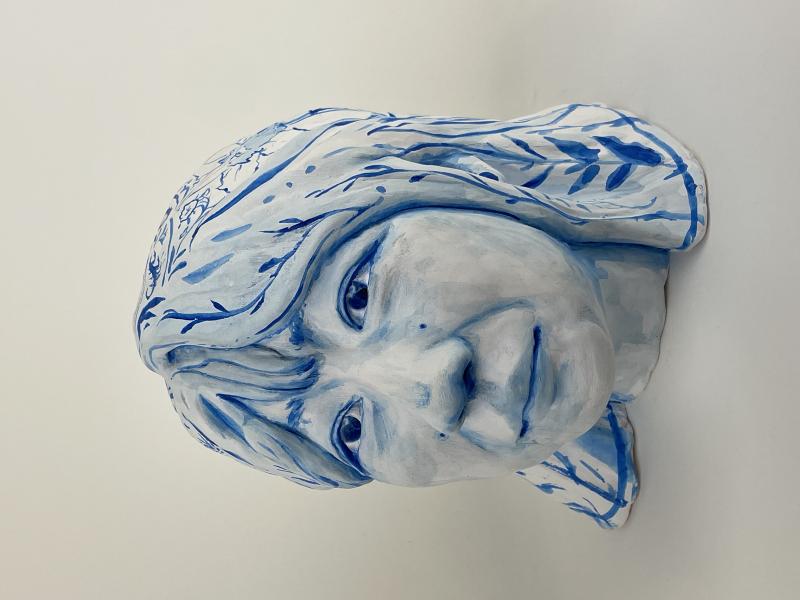 A ceramic bust of a person's head and shoulders, painted in shades of blue and white. The face has a calm expression with detailed features, and the hair is adorned with intricate blue patterns resembling leaves and branches.