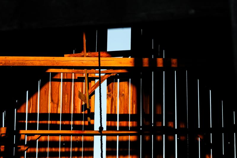 A close-up view of a wooden structure with vertical slats, illuminated by sunlight. The light creates strong shadows and highlights the texture of the wood. The background is partially visible through the gaps in the slats