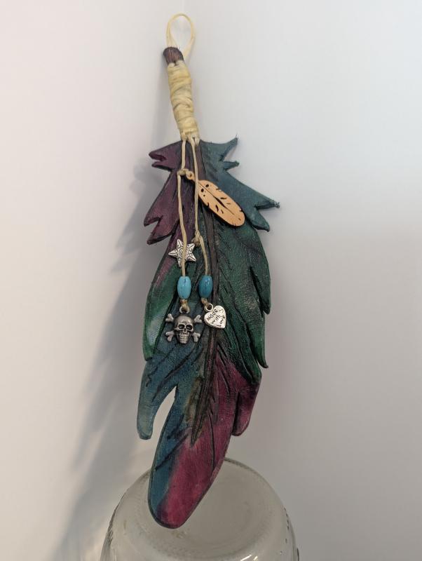 A decorative feather made of leather, painted in shades of green, blue, and purple. The feather is adorned with small charms including a silver star, turquoise beads, a small silver heart with an engraved design, and a wooden tag shaped like a smaller feather.