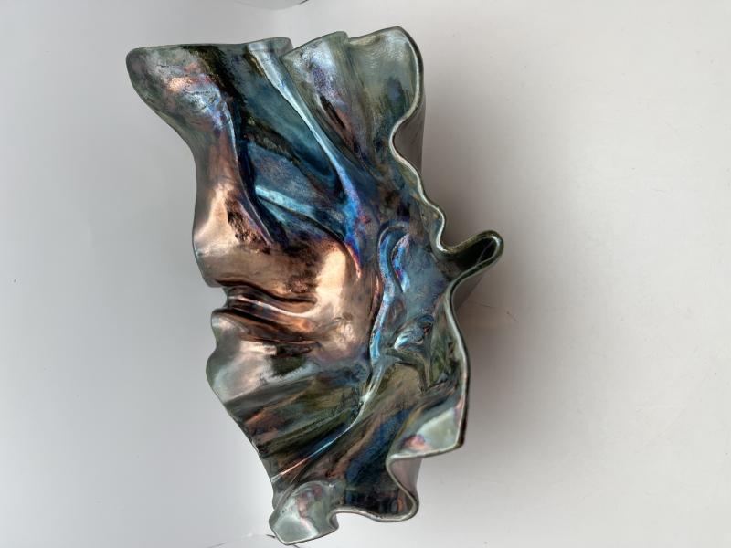 A metallic, abstract sculpture with an irregular, flowing shape. The surface has a reflective quality with hues of blue, pink, and silver.