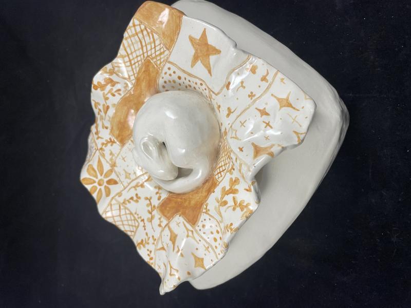 A ceramic sculpture featuring a central, smooth, white animal surrounded by an intricately patterned, orange and white square base with wavy edges. The base has various geometric and floral designs