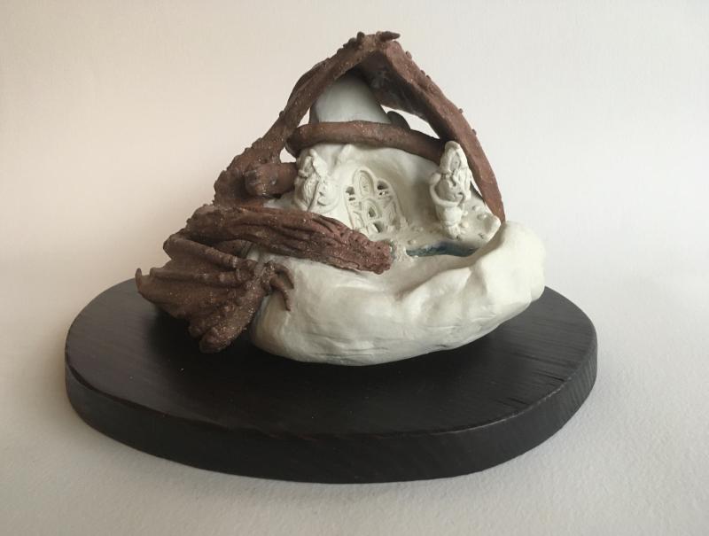 A small, intricate sculpture placed on a dark oval base. The sculpture appears to be made of clay and depicts a scene with detailed figures and structures inside what looks like a cave or grotto.