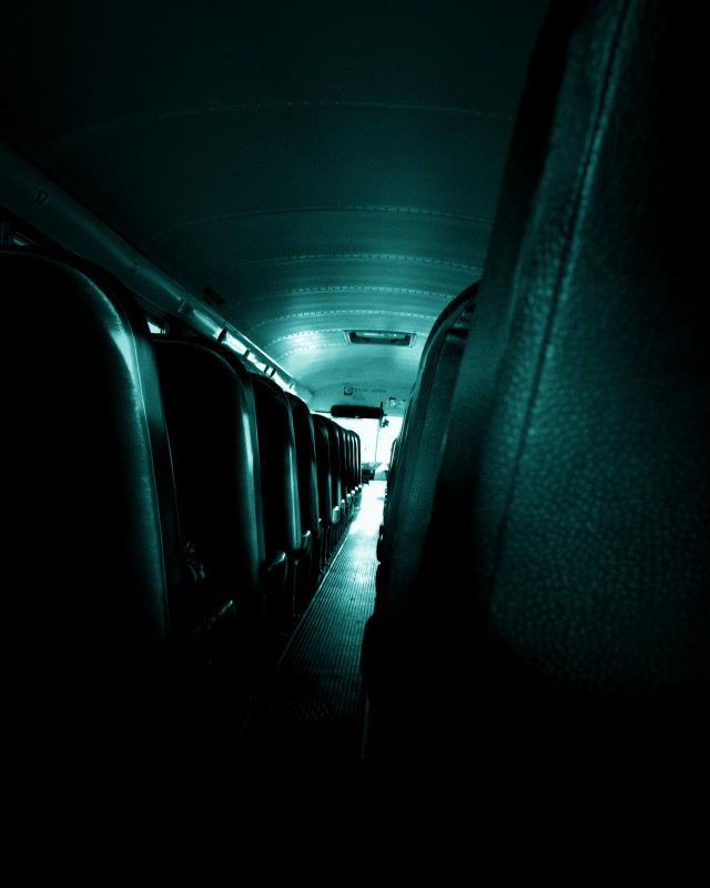 The interior of an empty bus, viewed from the back towards the front. The seats are arranged in rows on either side of a central aisle. The lighting is dim, casting a blue-green hue over the scene.