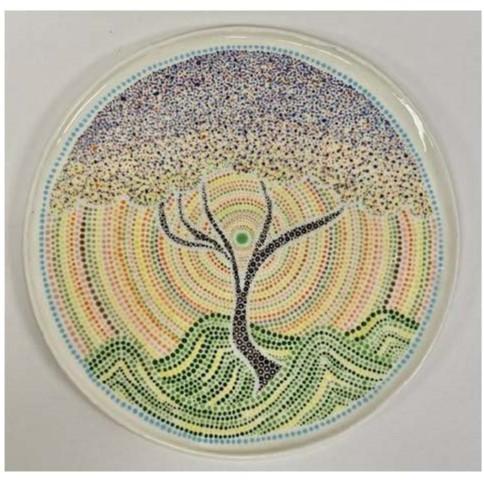 A circular artwork featuring a tree at the center, created using a pointillism technique. The tree has a black trunk and branches, with foliage depicted in various shades of blue, purple, and white dots. 
