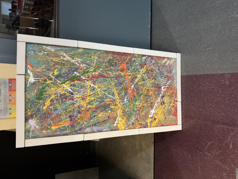 A rectangular abstract painting with vibrant splashes and streaks of various colors, including yellow, red, green, purple, and white. 