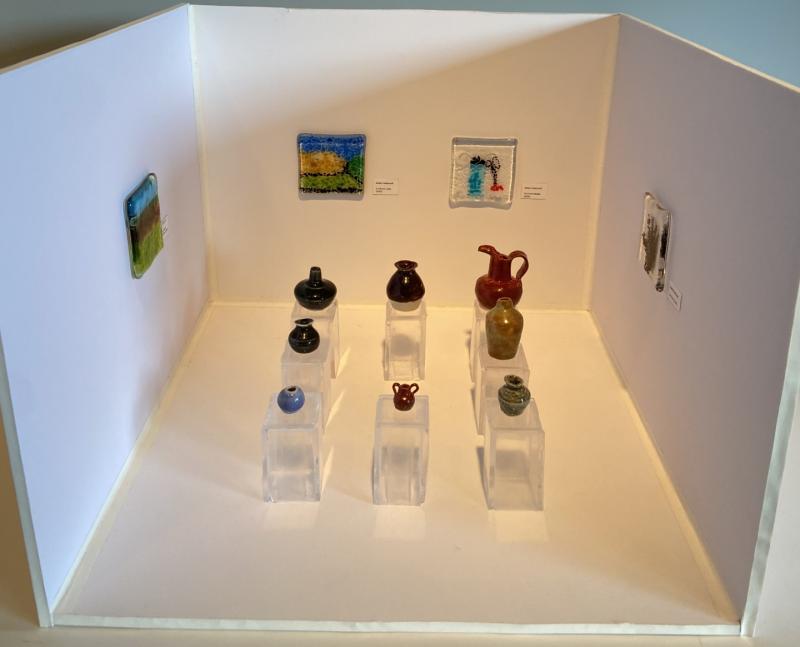 A miniature art gallery display featuring small pottery pieces and framed paintings. The display includes nine pottery items of various shapes and colors, placed on transparent pedestals. Three small framed paintings are mounted on the back wall, and two more are on the side walls