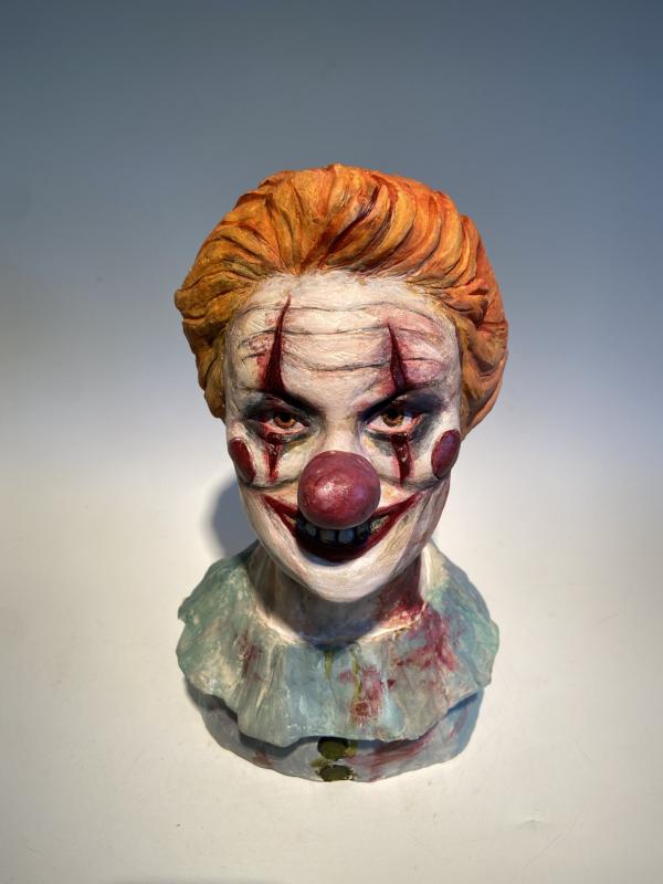 A sculpted bust of a sinister clown with exaggerated facial features, including a red nose, darkened eyes, and a wide, eerie grin revealing blackened teeth. The clown's face is painted white with red markings, and it wears a tattered, bloodstained ruffled collar. Its orange hair is slicked back, adding to its unsettling expression.