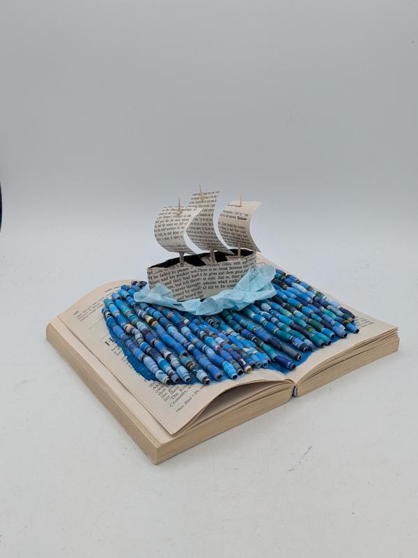 The artwork features an open book with a three-dimensional paper ship made from printed text pages sailing on a sea crafted from rolled blue paper beads. The ship has three sails and is positioned as if emerging from the book's pages.