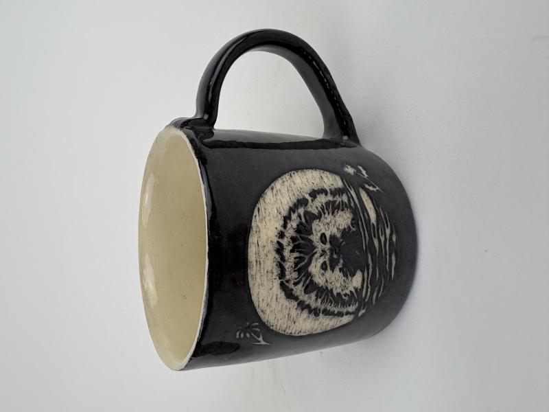 The artwork is a black ceramic mug with a white interior, featuring an etched design of an owl on one side. The owl is depicted in a detailed and textured style, with its face and feathers clearly visible. 