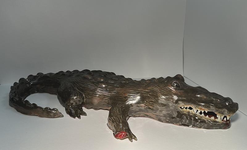 The artwork is a realistic sculpture of a crocodile displayed against a plain background. The sculpture is detailed, showing the textured skin, sharp teeth, and claws of the crocodile. 