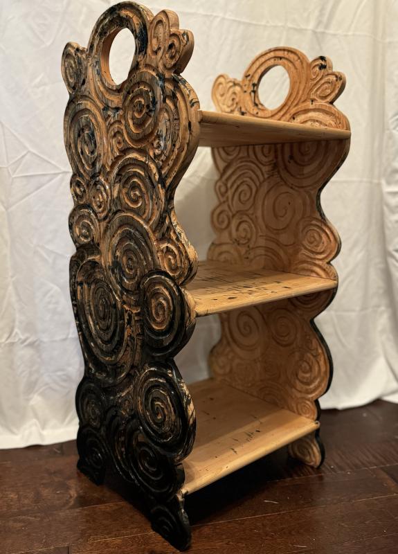 The artwork is a uniquely designed wooden shelf with three tiers. The sides of the shelf feature intricate, swirling patterns that resemble clouds or waves, with a mix of natural wood and black paint. 