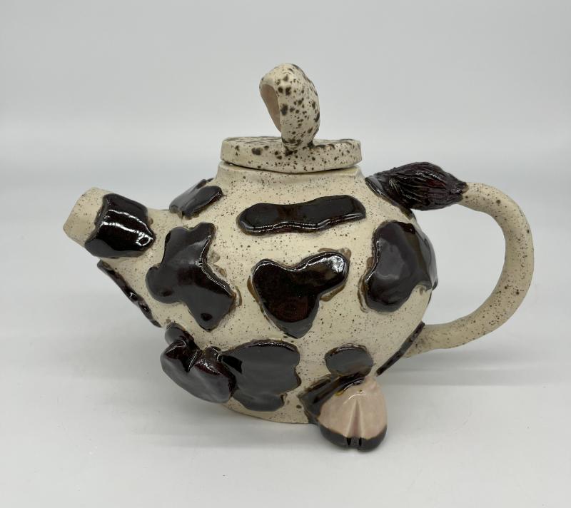 The artwork is a ceramic teapot designed to resemble a cow. The teapot is primarily white with black spots, mimicking the pattern of a cow's hide. The spout and handle are shaped like a cow's head and tail, respectively. 