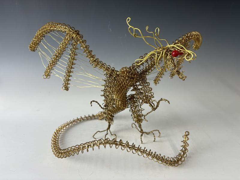The image shows a detailed sculpture of a dragon made from intricately twisted and coiled wire. The dragon has large wings, a long tail, and an expressive face with what appears to be a red bead for an eye. 