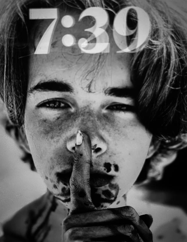 Black-and-white portrait of a child with messy hair and dirt-stained skin, holding a finger to their lips in a gesture of silence. The time "7:39" is overlaid at the top.