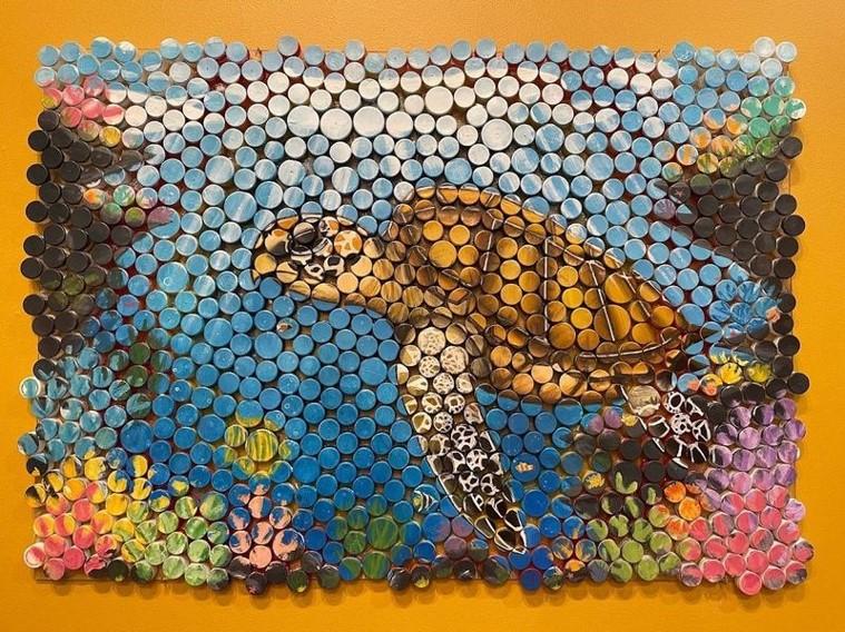 “Recycled Ocean” is a mixed media painting of a Pacific Loggerhead Turtle swimming in a coral reef clearing. The artist used plastic bottle caps to create a mosaic of this scene. 