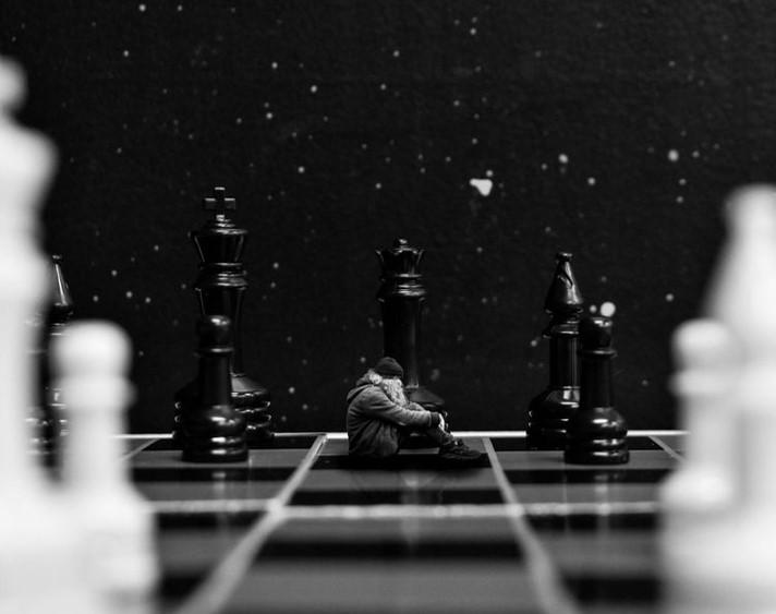 A black and white photograph of the artist sitting on a chessboard amongst the game pieces.