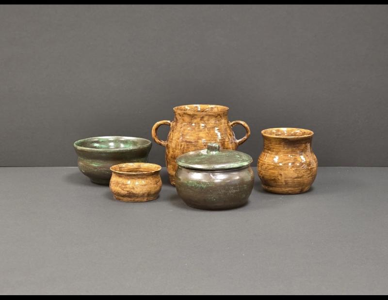 Five handmade ceramic vessels displayed against a dark background, including bowls and handled jars, finished in earthy brown and deep green glazes with subtle texture and softly rounded forms.